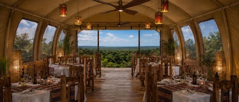 A tented dining area with tables set for a meal, a ceiling fan, and large windows offering a view of the landscape.
