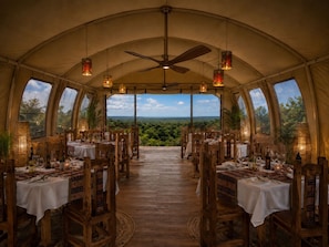 A tented dining area with tables set for a meal, a ceiling fan, and large windows offering a view of the landscape.