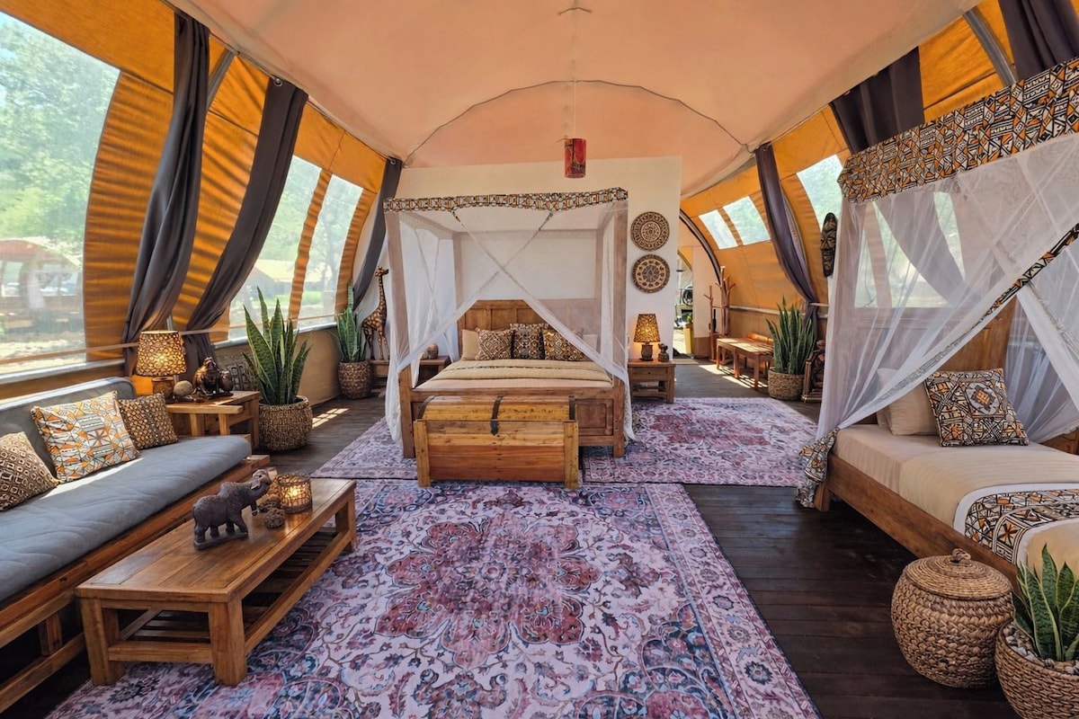 A tented accommodation with a central bed, two sofas, a dining area, and a view of the outdoors.