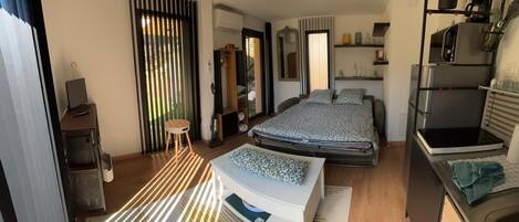 A modern hotel room with a bed, a desk, a chair, a wardrobe, a mirror, a kitchen area, and a sliding door.