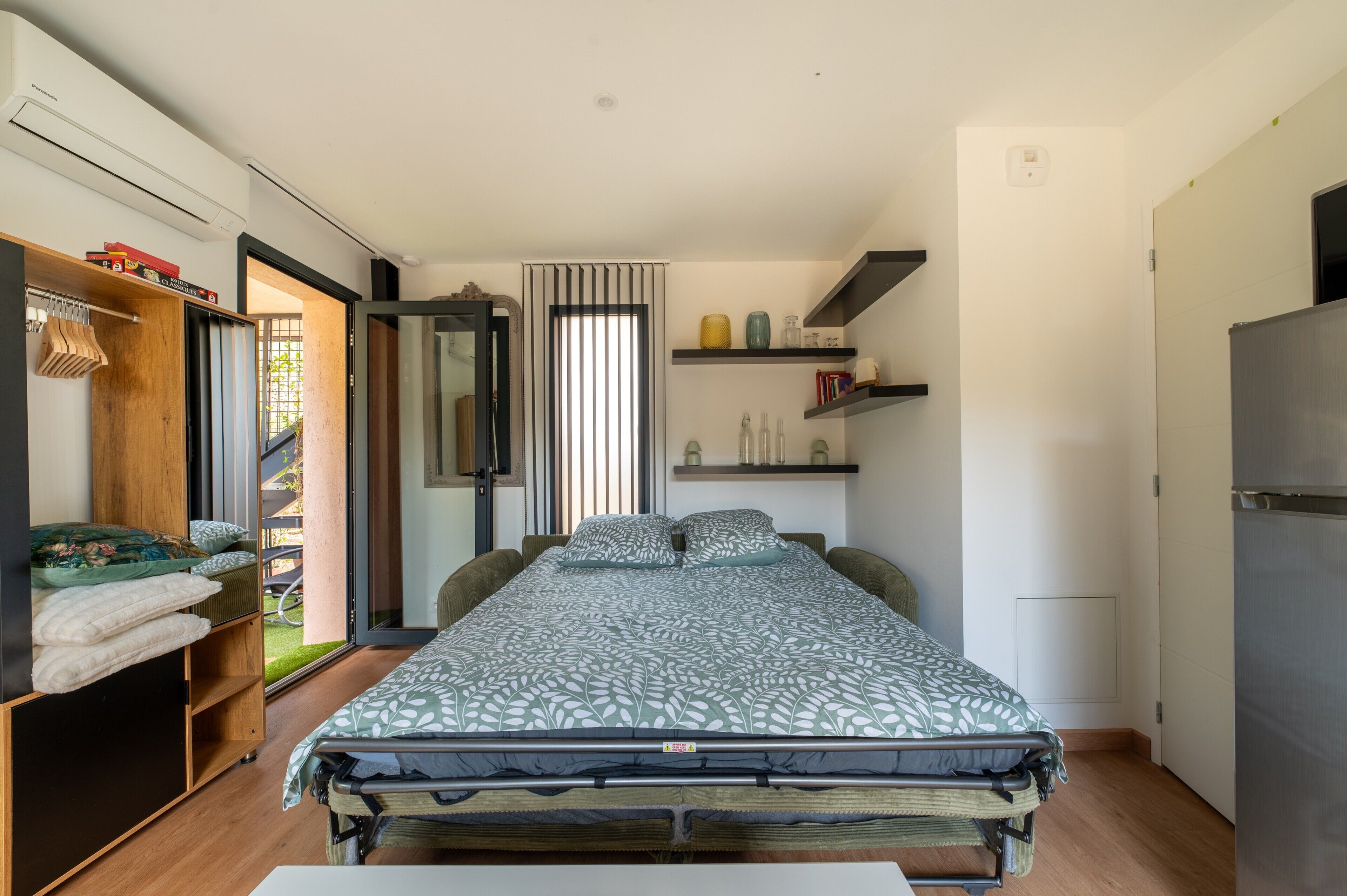 Un dormitorio compacto con cama, armario, estante con objetos decorativos y puerta que da a un espacio exterior.