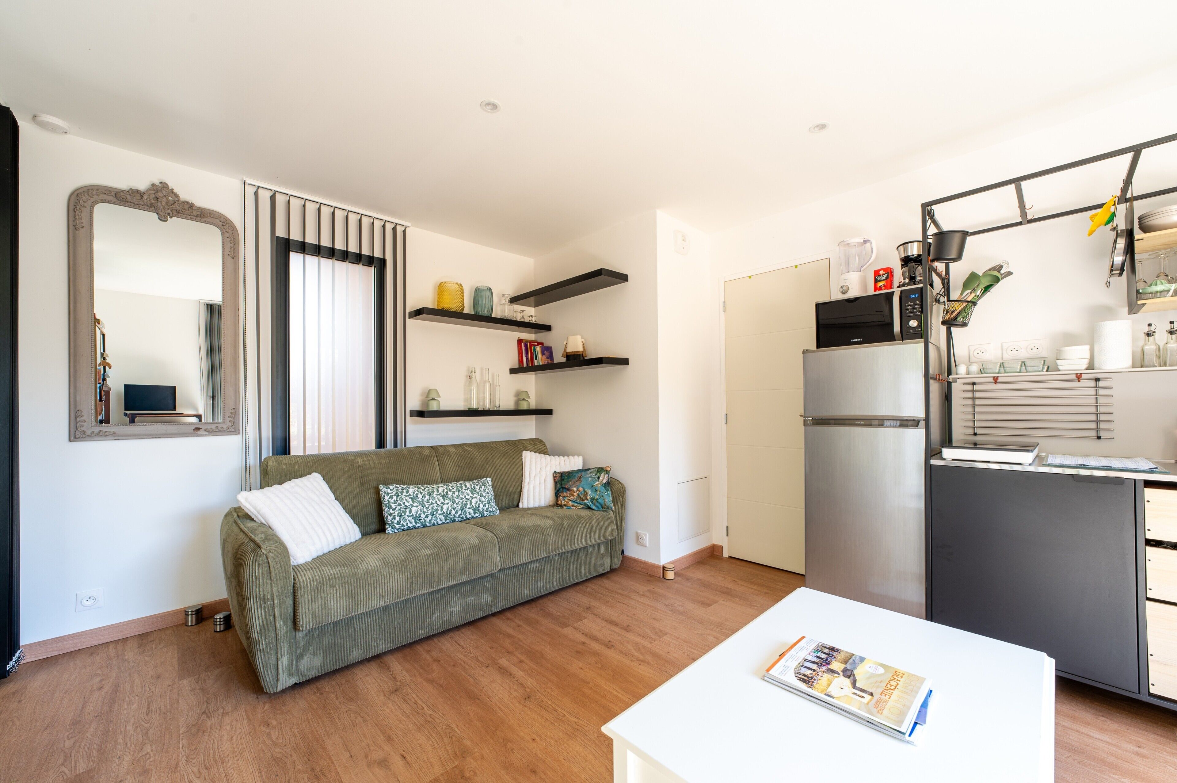 A compact living space with a sofa, a dining table, a kitchen area with a refrigerator, and a mirror.