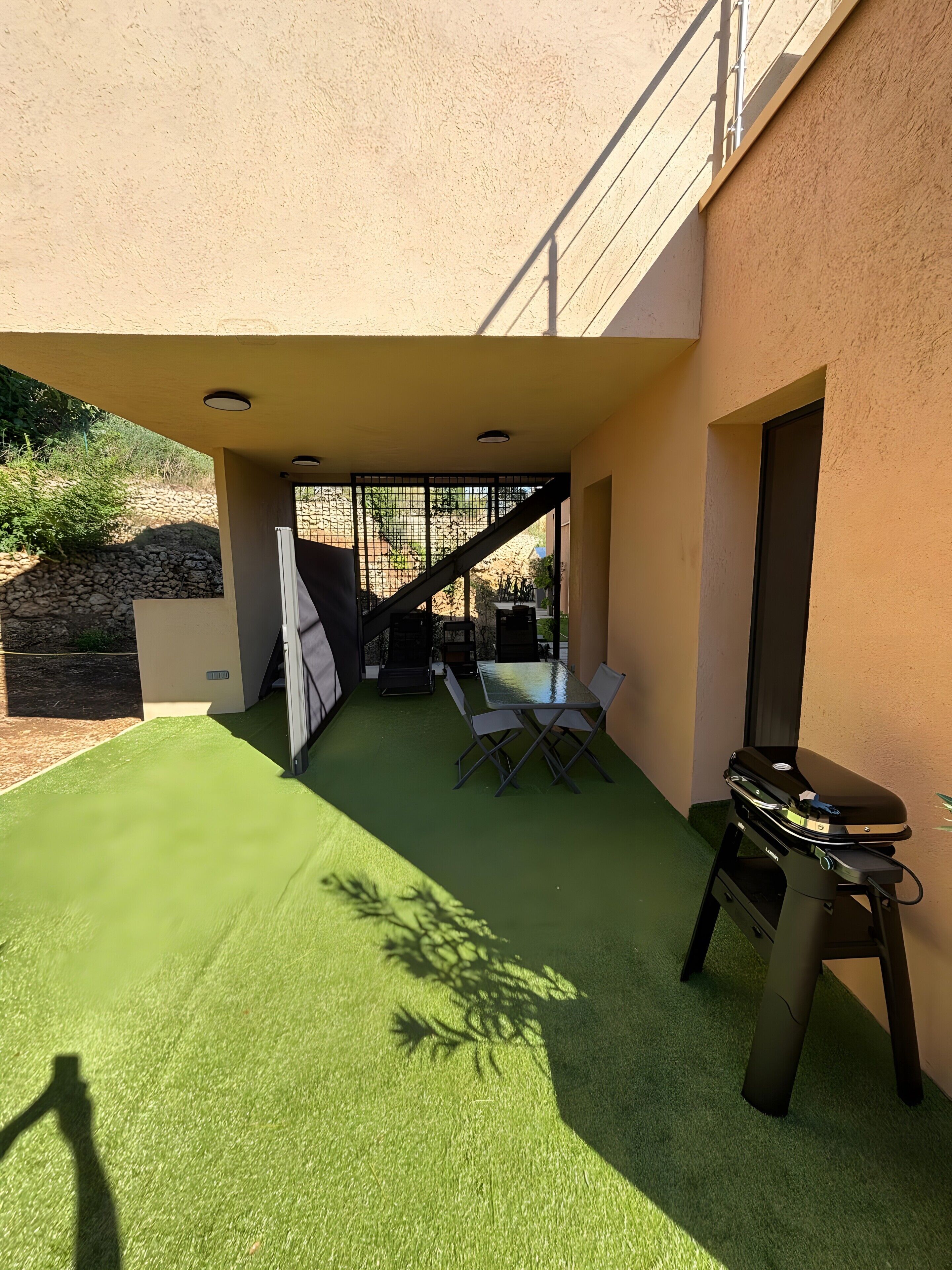 Une terrasse extérieure avec un revêtement de gazon synthétique, une table et des chaises, un barbecue et un mur de pierres en arrière-plan.