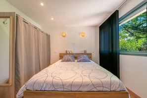 Ein Schlafzimmer mit einem Bett, zwei Wandleuchten, einem Fenster mit Vorhängen und Blick auf Grünflächen.