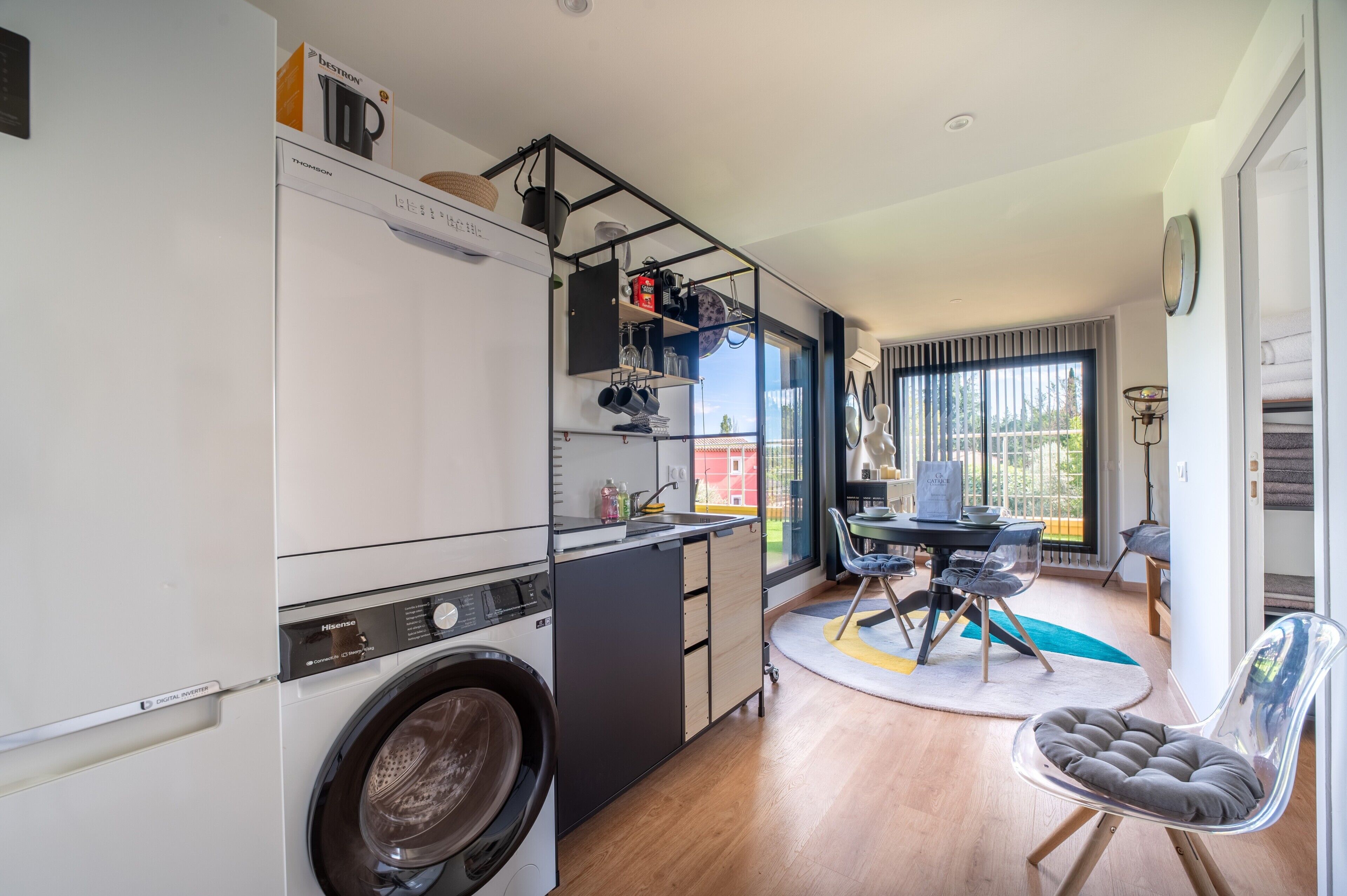 Une cuisine compacte équipée d’un lave-linge, un coin repas avec une table et des chaises, et une vue par la fenêtre.