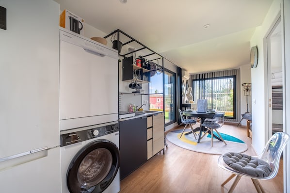A compact kitchen with a washing machine, a dining area with a table and chairs, and a view through the window.