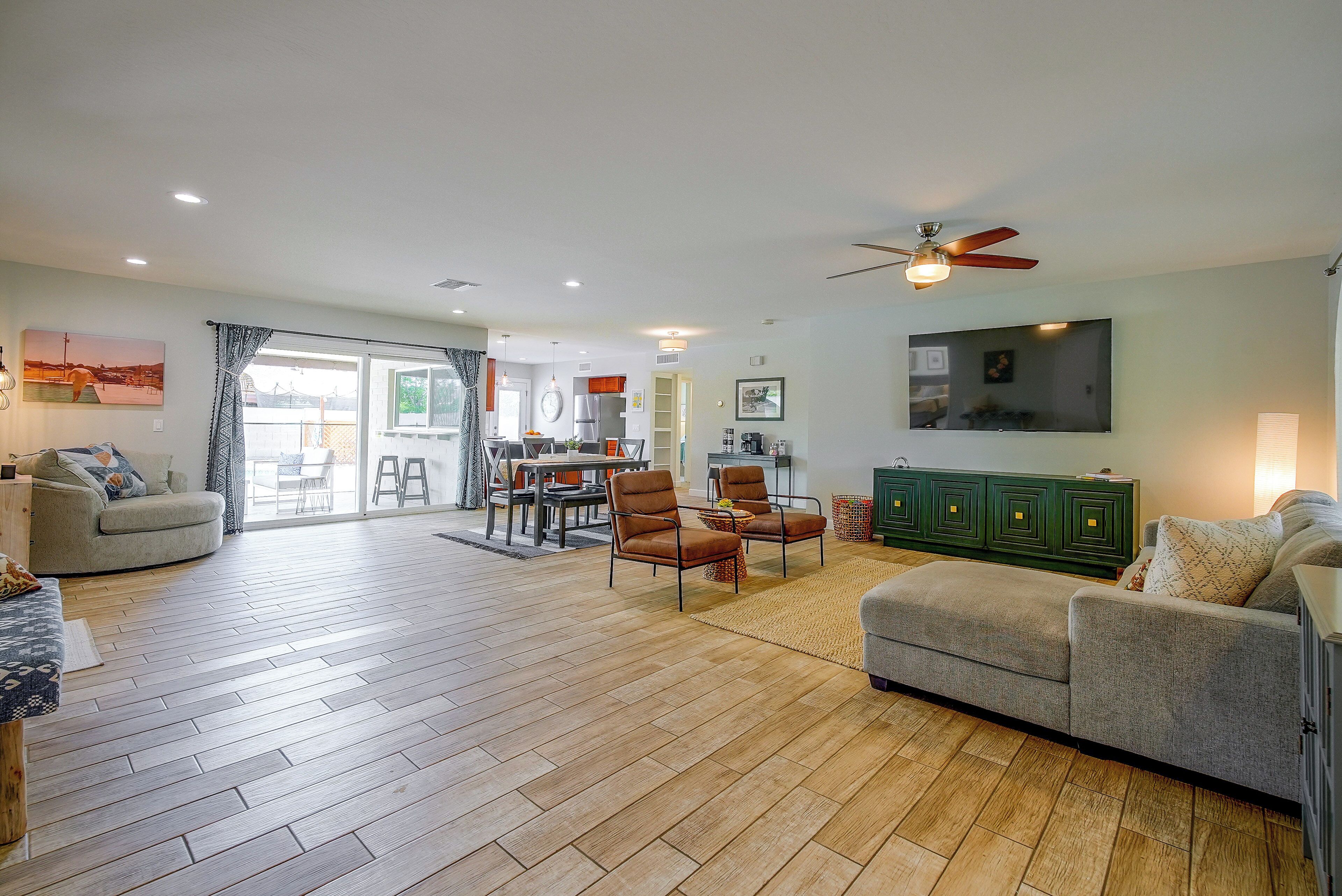 A spacious living room with a dining area, a kitchen in the background, and a large TV mounted on the wall.