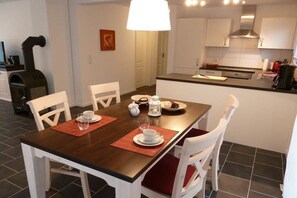 Un comedor con una mesa de madera, sillas blancas y una cocina al fondo.