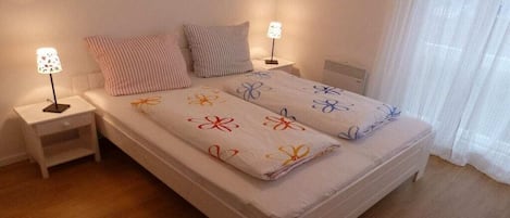 Un lit simple avec une couvre-lit à motifs floraux, deux lampes de chevet, une table de chevet et une fenêtre avec des rideaux légers.