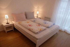 Una cama individual con colcha estampada, dos lámparas de noche, una mesita auxiliar y una ventana con cortinas traslúcidas.