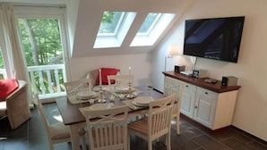Uma sala de jantar com mesa posta para quatro pessoas, televisão na parede e janela com vista para área verde.