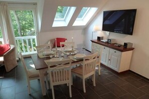 A dining area with a table set for four, a television mounted on the wall, and a window with a view of greenery. - Apartment 4.3 – Holiday apartment in Graal-Müritz (Graal-Müritz)
