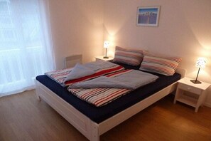 Habitación con una cama individual, cabecera blanca, dos mesitas de noche con lámparas, una ventana con cortinas traslúcidas y un cuadro en la pared.