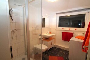 Un baño con cabina de ducha de vidrio, inodoro blanco, lavabo y una ventana sobre la bañera.
