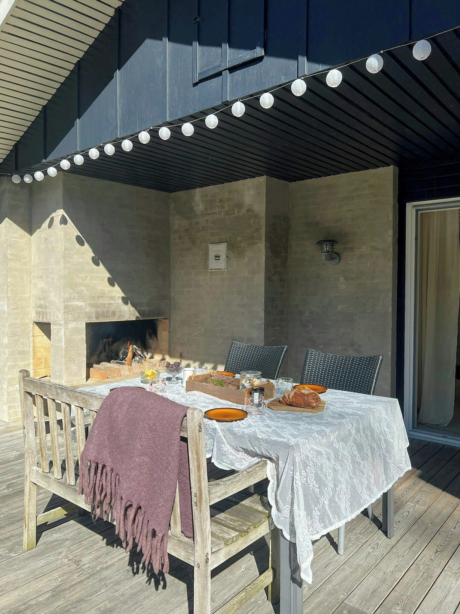 An outdoor dining area with a table set for two, surrounded by chairs, a wooden bench with a blanket, and string lights overhead.