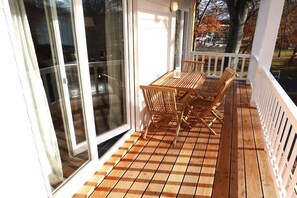 Una terraza de madera con mesa y sillas, baranda blanca y puerta de vidrio que da a un patio.