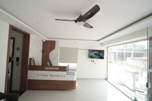Un comptoir d’accueil doté d’un ventilateur de plafond, d’un panneau indiquant les heures d’arrivée et de départ, et d’une grande fenêtre donnant sur l’extérieur.