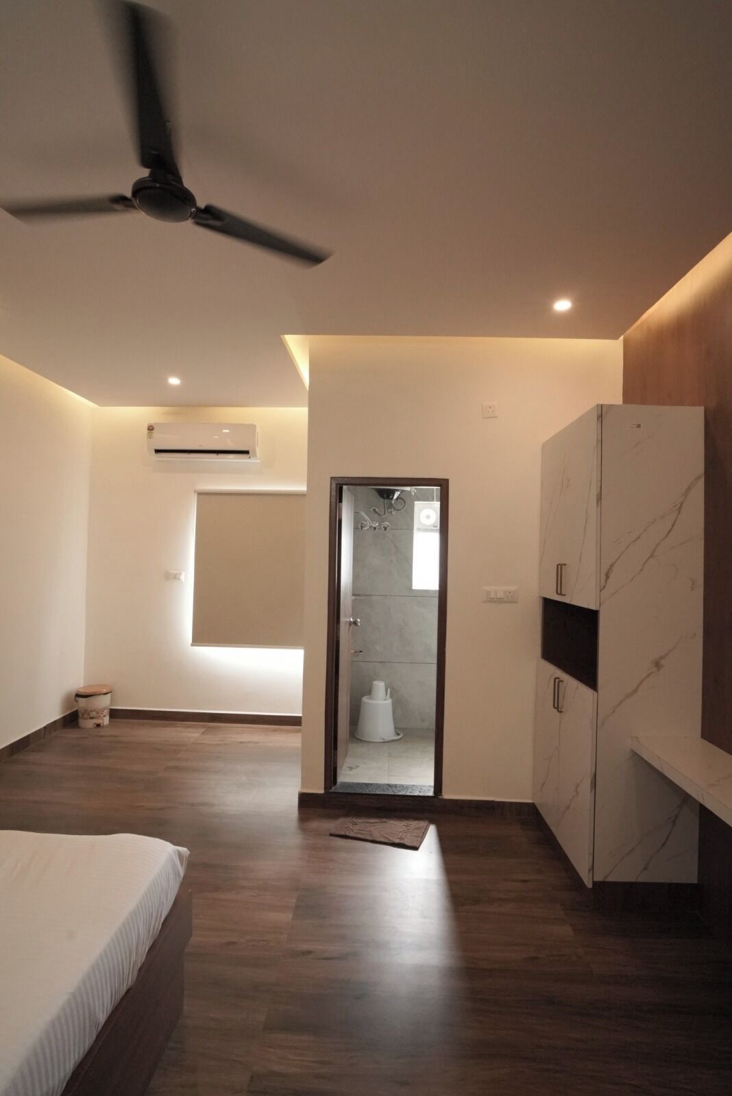 Un dormitorio moderno con cama, ventilador de techo, equipo de aire acondicionado y vista al baño a través de una puerta de cristal.