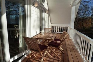 Una terraza techada al aire libre con una mesa y sillas, barandal blanco y vista a árboles.