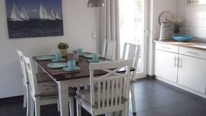 Uma sala de jantar com mesa posta para quatro pessoas, cadeiras brancas, um quadro com veleiros na parede e uma área de cozinha com armários e pia.