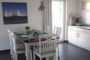 Un comedor con una mesa dispuesta para cuatro, sillas blancas, un cuadro de veleros en la pared y una zona de cocina con armarios y un fregadero.