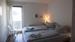 Quarto com duas camas, janela com cortinas, mesa de cabeceira com abajur e uma cadeira.