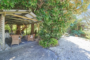 A covered outdoor seating area with wicker furniture, surrounded by lush greenery and a gravel path. - Bergonia by Hyams Beach Seaside Cottages | Romantic, iconic heritage cottage (Hyams Beach)