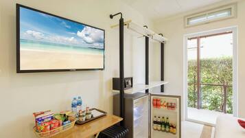 A room with a television displaying a beach scene, a small refrigerator stocked with beverages, a table with snacks and a basket, and a window with a view of greenery.