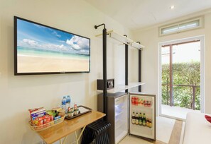 A room with a television displaying a beach scene, a small refrigerator stocked with beverages, a table with snacks and a basket, and a window with a view of greenery. - Villa 21D - GOLF Room (ตำบล เกาะช้าง)