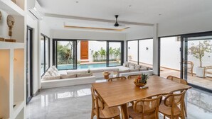 Een moderne eetruimte met een houten tafel en stoelen, een bank met kussens en uitzicht op een zwembad door grote ramen.