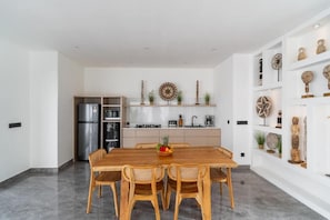 A modern kitchen with white cabinetry, a stainless steel refrigerator, and a dining area with a wooden table and wicker chairs. - Majys 1 Villa by Tasvan (Badung)