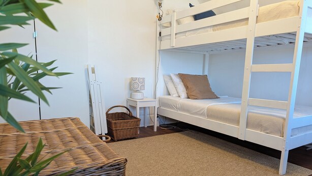 A bunk bed with white bedding, a small table with a lamp, a wicker basket, and a plant. - Vrbo Property (Staten Island)