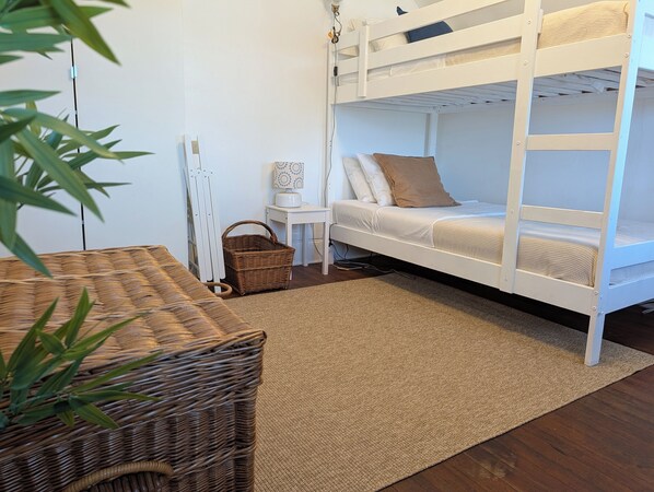 Una cama litera con ropa de cama blanca, una mesita con una lámpara, una cesta de mimbre y una planta.