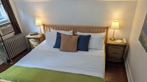 A bed with a wooden frame, white bedding, and a green throw. There are two bedside lamps, a radiator, and a small table with a lamp and a phone. - Vrbo Property (Staten Island)