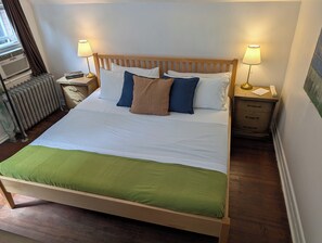 Una cama con estructura de madera, ropa de cama blanca y un plaid verde. Hay dos lámparas de noche, un radiador y una mesita con una lámpara y un teléfono.