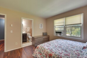 Een slaapkamer met een houten dressoir, een bed met bloemenbedrukking, een raam met zonwering en uitzicht op bomen.
