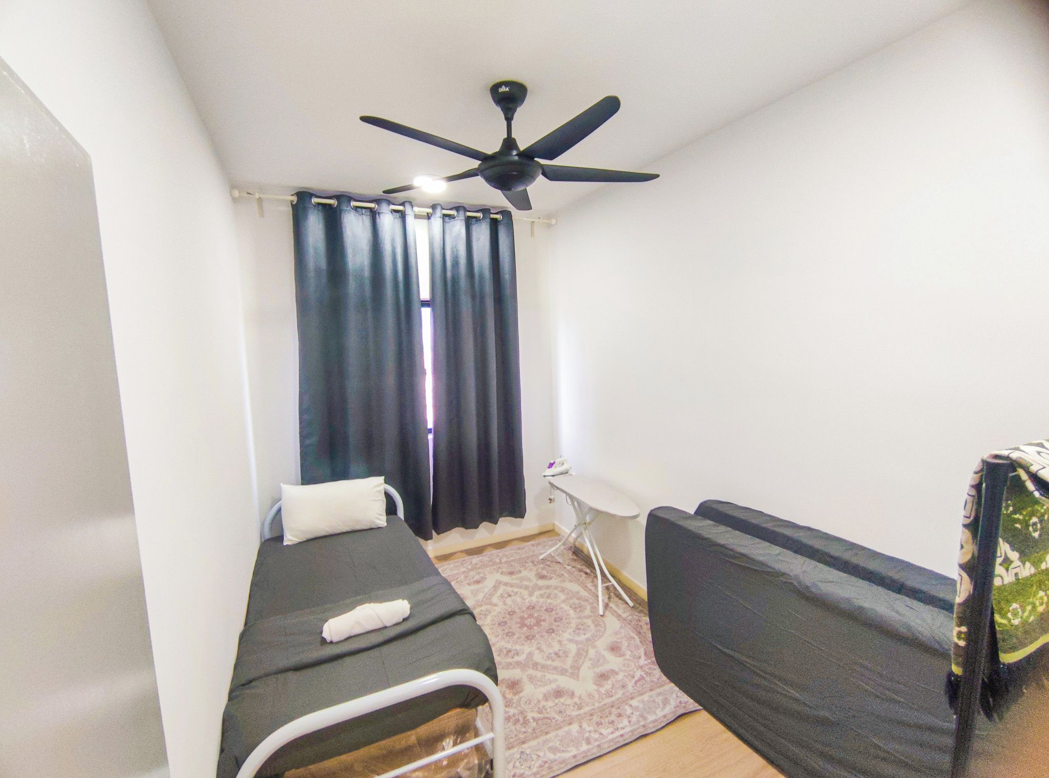 Une petite chambre avec un lit simple, un ventilateur de plafond, une table de chevet, une chaise et un tapis à motifs.