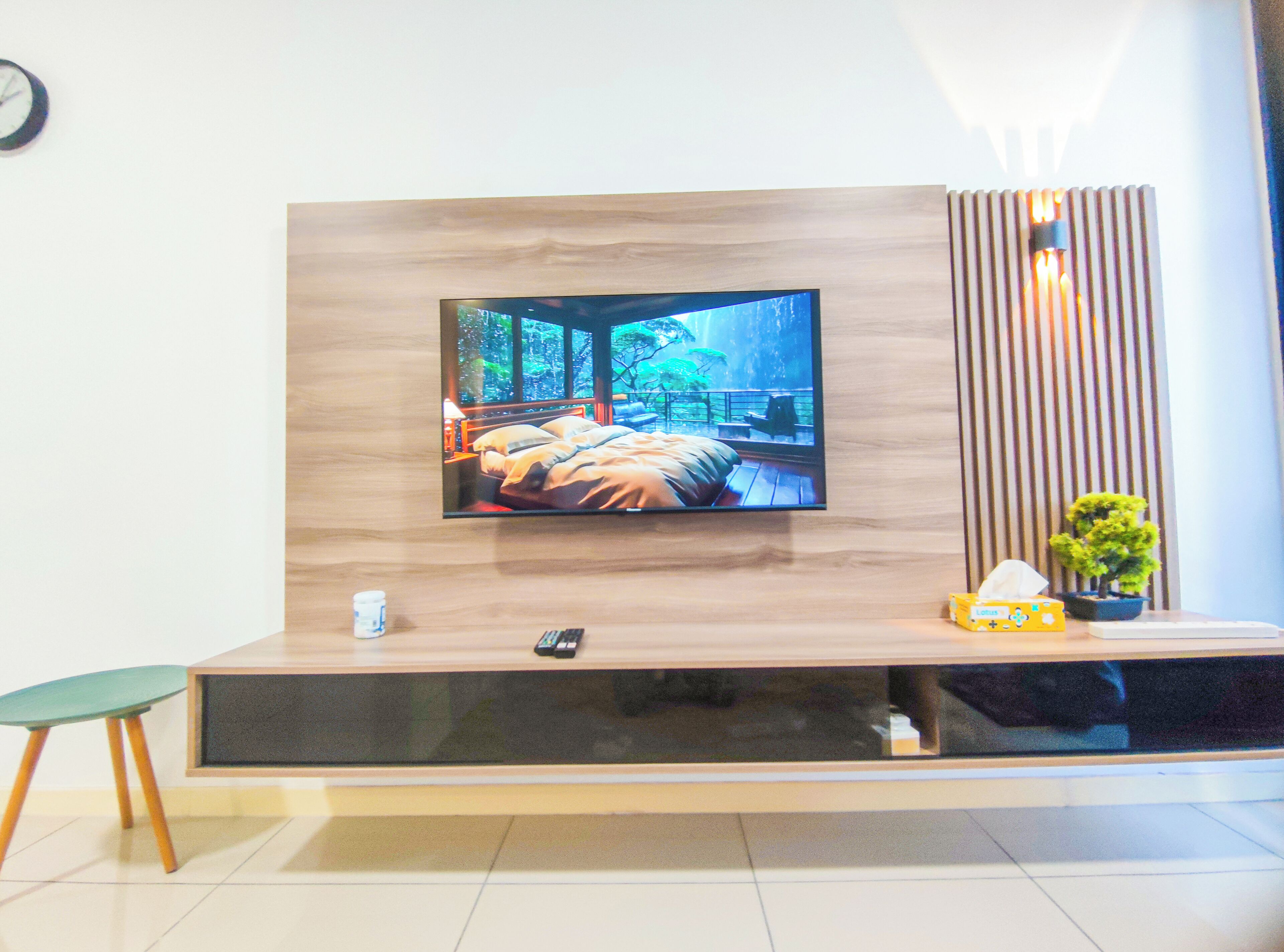 A modern living room with a flat-screen TV mounted on a wooden wall, a small table with a remote control, a green chair, and a potted plant.