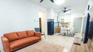 Una sala de estar con sofá, mesa de centro y televisor. Se observa una cocina al fondo con armarios y comedor.