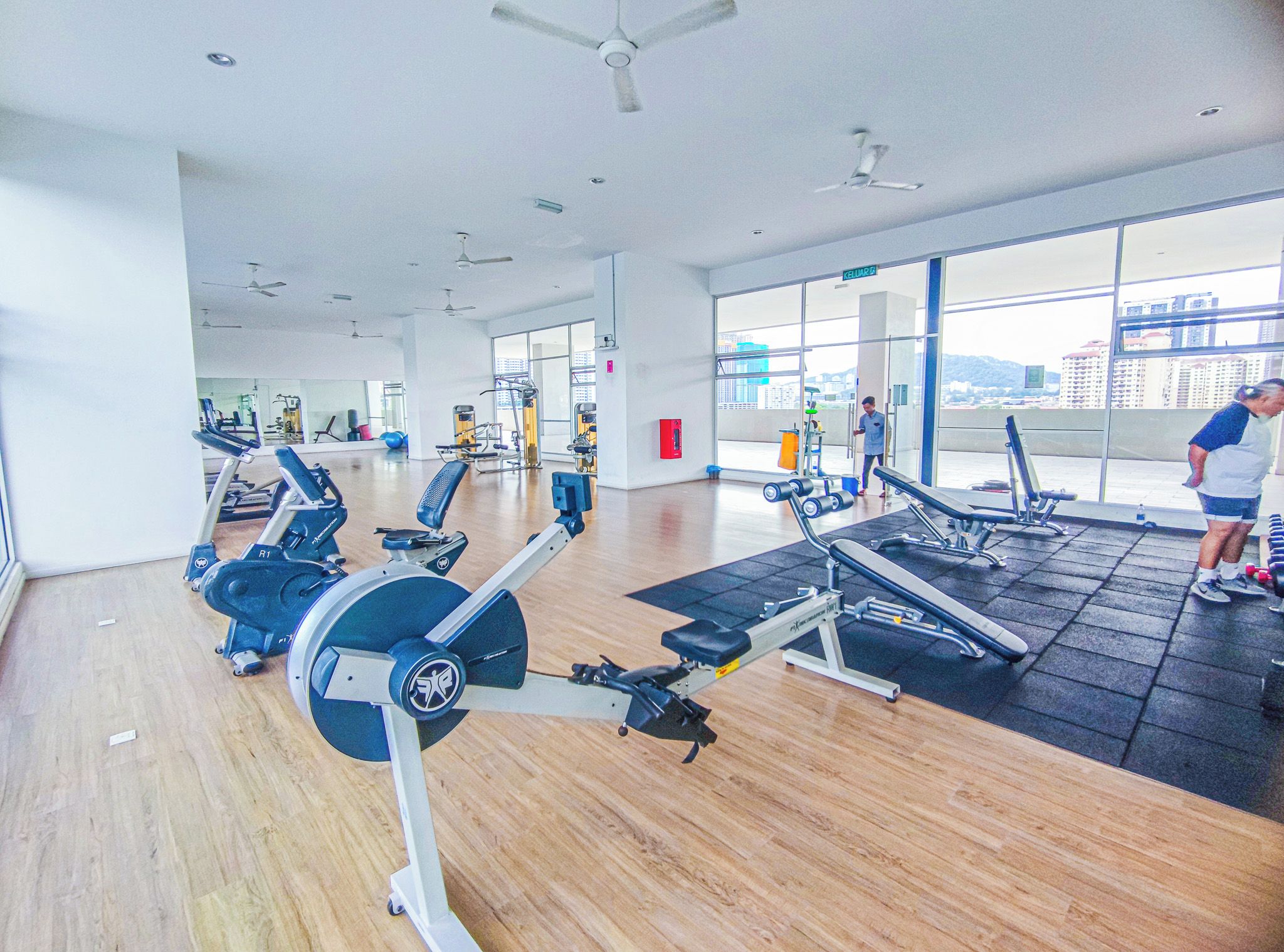 Ett gym med konditionsmaskiner, träningsbänkar och ett stort fönster med utsikt över staden.