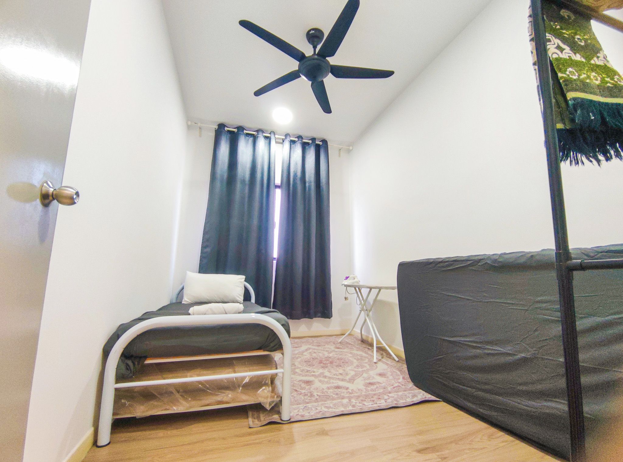 Habitación pequeña y compacta con cama, ventilador de techo, una mesita y ventana con cortinas.