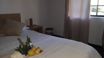 Una cama bien hecha con una colcha blanca, una toalla enrollada y una pequeña planta con flores amarillas.