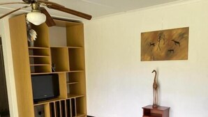Un salon avec un ventilateur de plafond, un téléviseur posé sur un meuble en bois, un canapé marron et un sol carrelé.