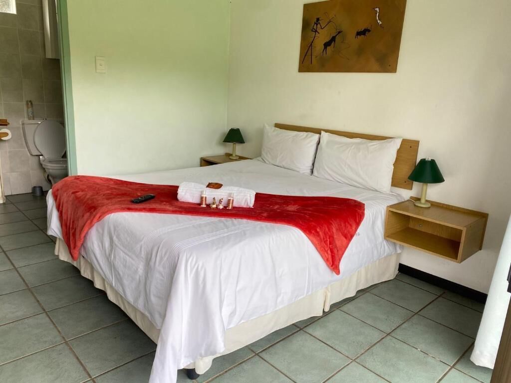 Une chambre avec un lit simple, une tête de lit en bois, du linge de lit blanc, un plaid rouge, des lampes de chevet et un tableau au mur.