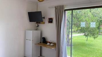 Una habitación con ventilador de techo, televisión sobre un mueble bajo, refrigerador pequeño, ventana con cortinas y vista a un área con césped.