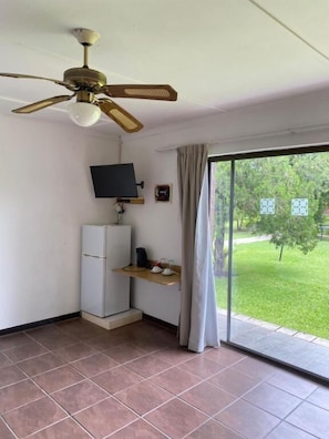 A room with a ceiling fan, a television on a stand, a small refrigerator, a window with curtains, and a view of a grassy area. - Thornycroft Lodge & Spa KuGompo City (KuGompo City)
