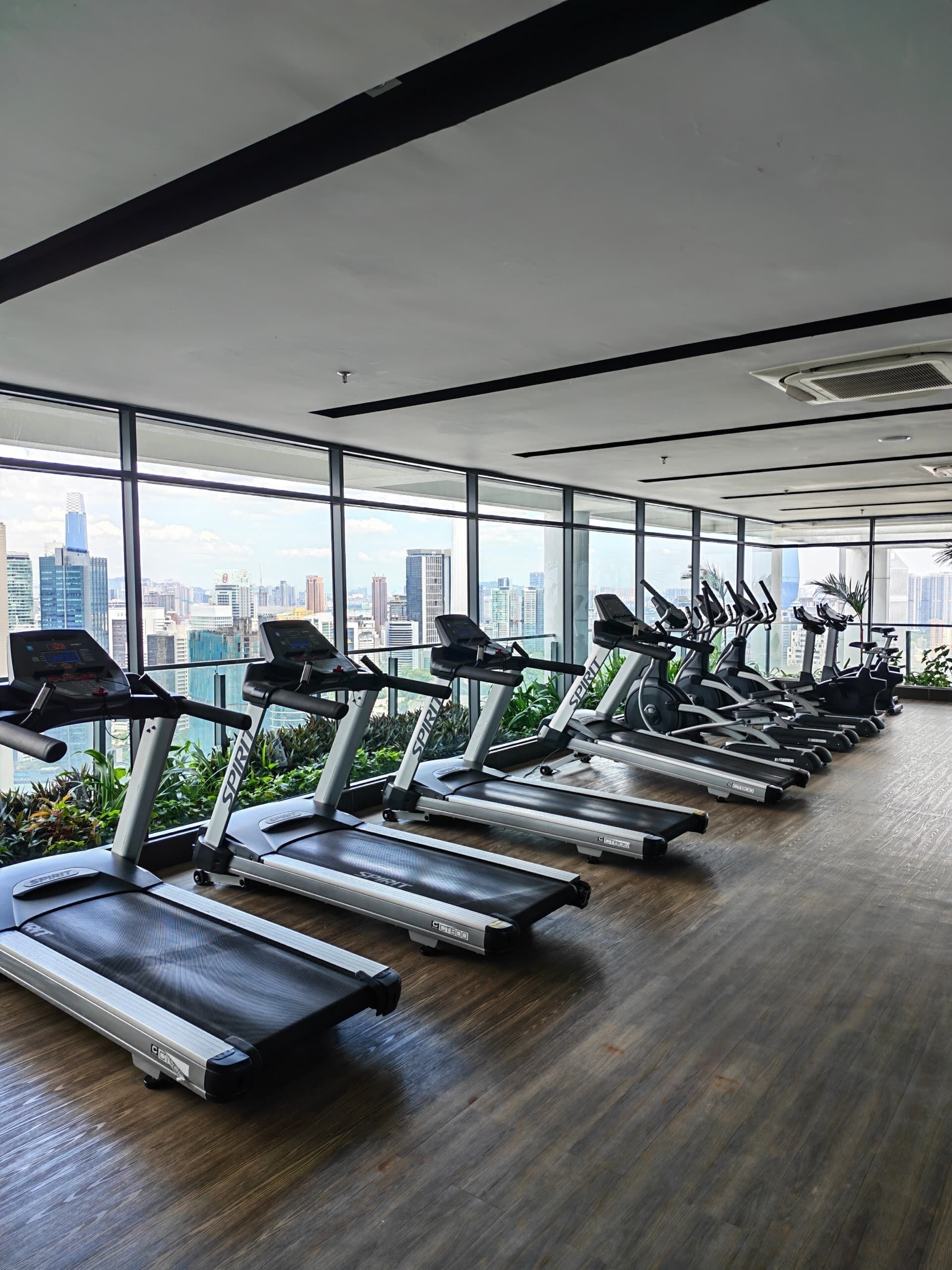 A gym with multiple treadmills aligned in rows, large windows offering a city view, and a wooden floor.