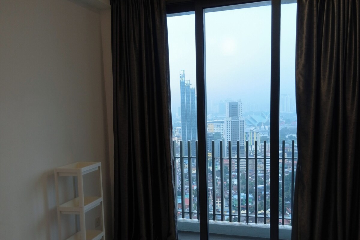 Ein Hotelzimmer mit Bett, weißem Regal und Blick auf eine Stadtlandschaft durch große Fenster mit dunklen Vorhängen.