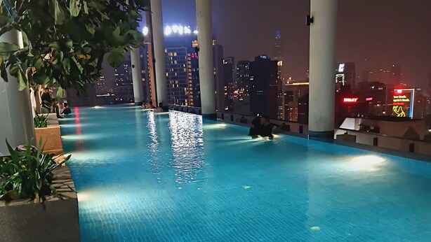 An indoor swimming pool with clear blue water, surrounded by columns and plants, with a cityscape view in the background. - Infinitum by colony  (Kuala Lumpur)