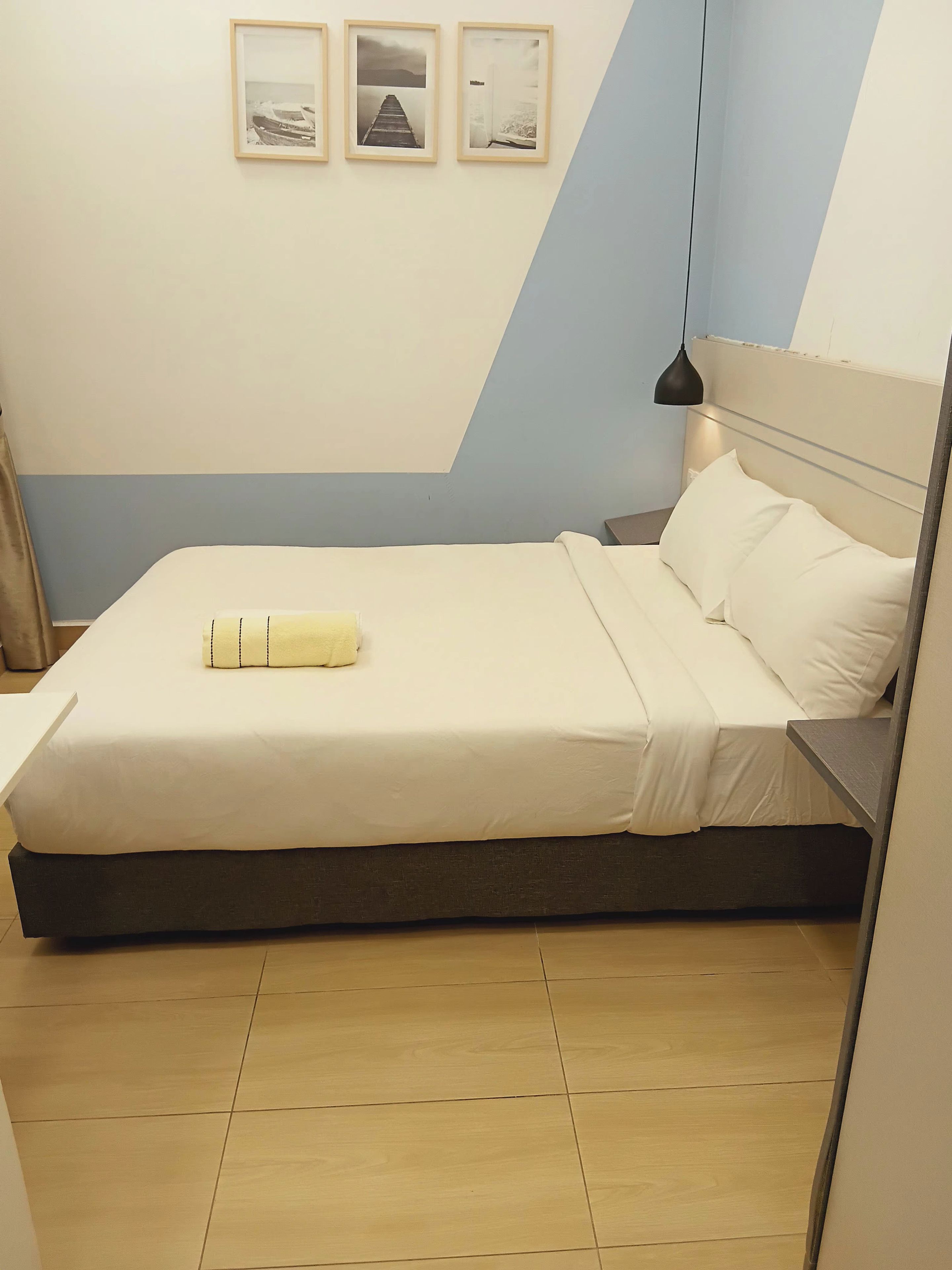 Una cama bien hecha con una colcha blanca y almohadas, una mesita de noche con una lámpara, y una pared con tres cuadros enmarcados.